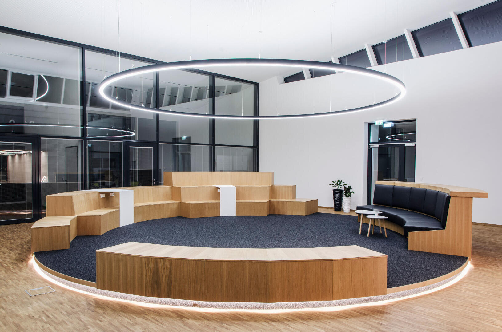 Une salle de réunion ronde et moderne avec des sièges en bois étagés, un banc noir rembourré, de petites tables et un grand plafonnier rond dans une pièce aux murs de verre et aux sols en bois.