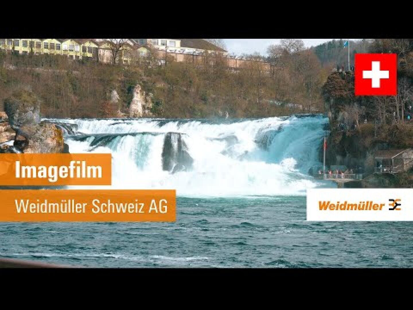Ein breiter Wasserfall fließt unter bewölktem Himmel, umgeben von Bäumen und Gebäuden. Oben rechts ist die Schweizer Flagge zu sehen, und der orangefarbene Text lautet Imagefilm Weidmüller Schweiz AG by Weidmüller.