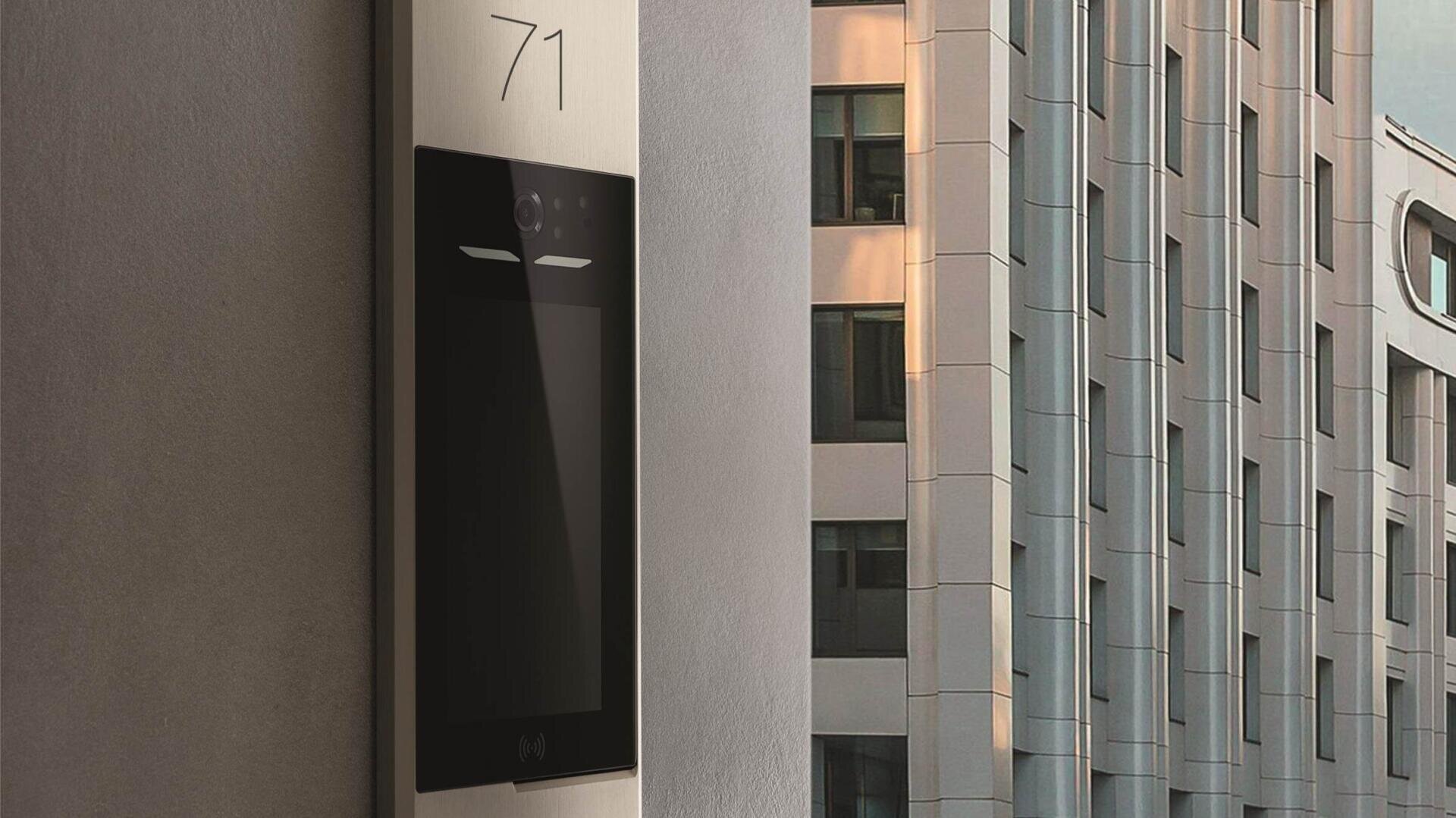 Ein modernes Gebäudeeingangstableau mit digitaler Gegensprechanlage und der Nummer 71 ist an einer Wand montiert. Im Hintergrund sind die hohen Fenster eines anderen Gebäudes zu sehen.