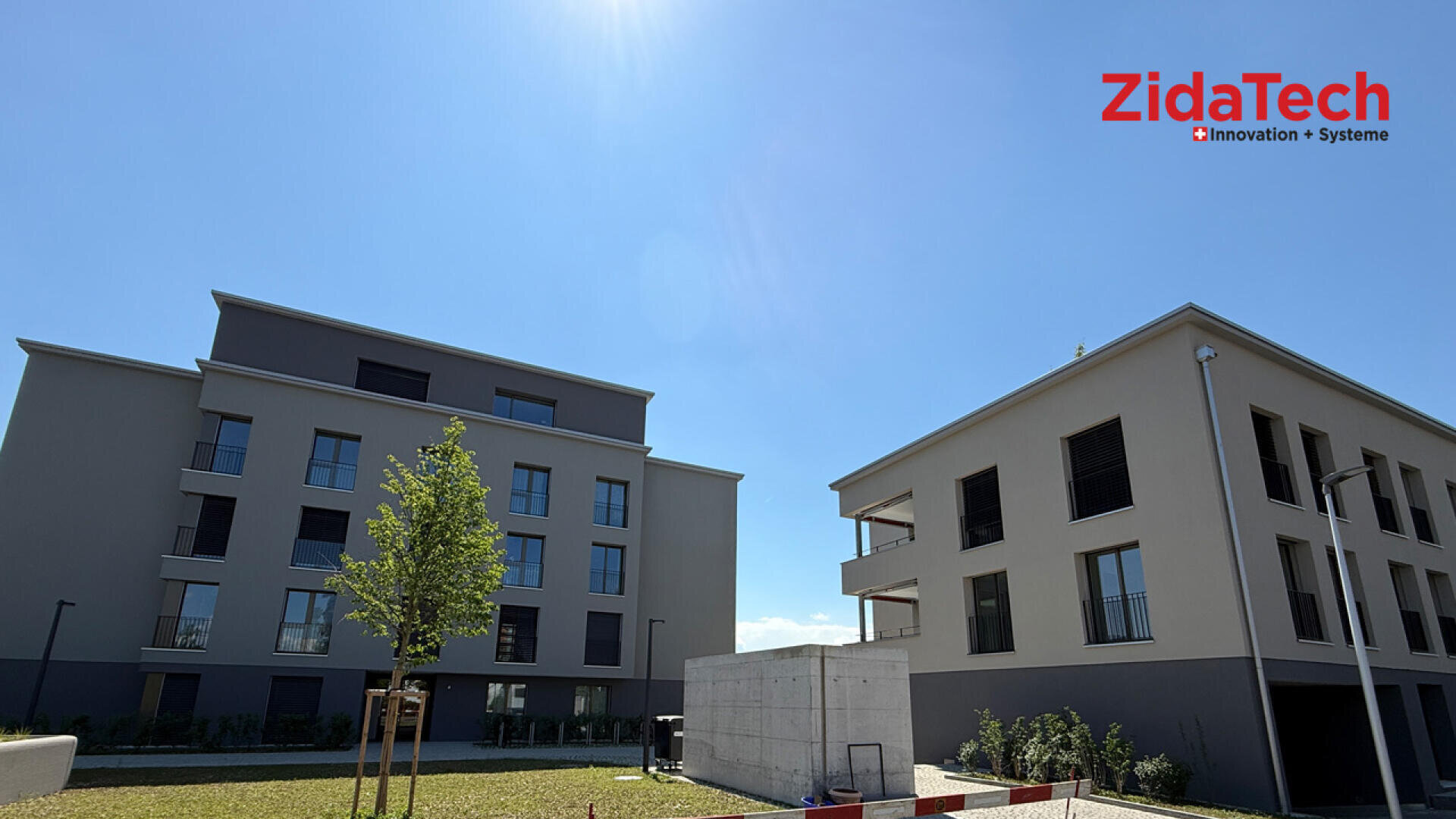 Zwei moderne Gebäude mit großen Fenstern erheben sich unter einem hellblauen Himmel. Sie sind durch einen jungen Baum und eine Rasenfläche voneinander getrennt. Das Logo von ZidaTech Innovation Systeme erscheint in der oberen rechten Ecke.