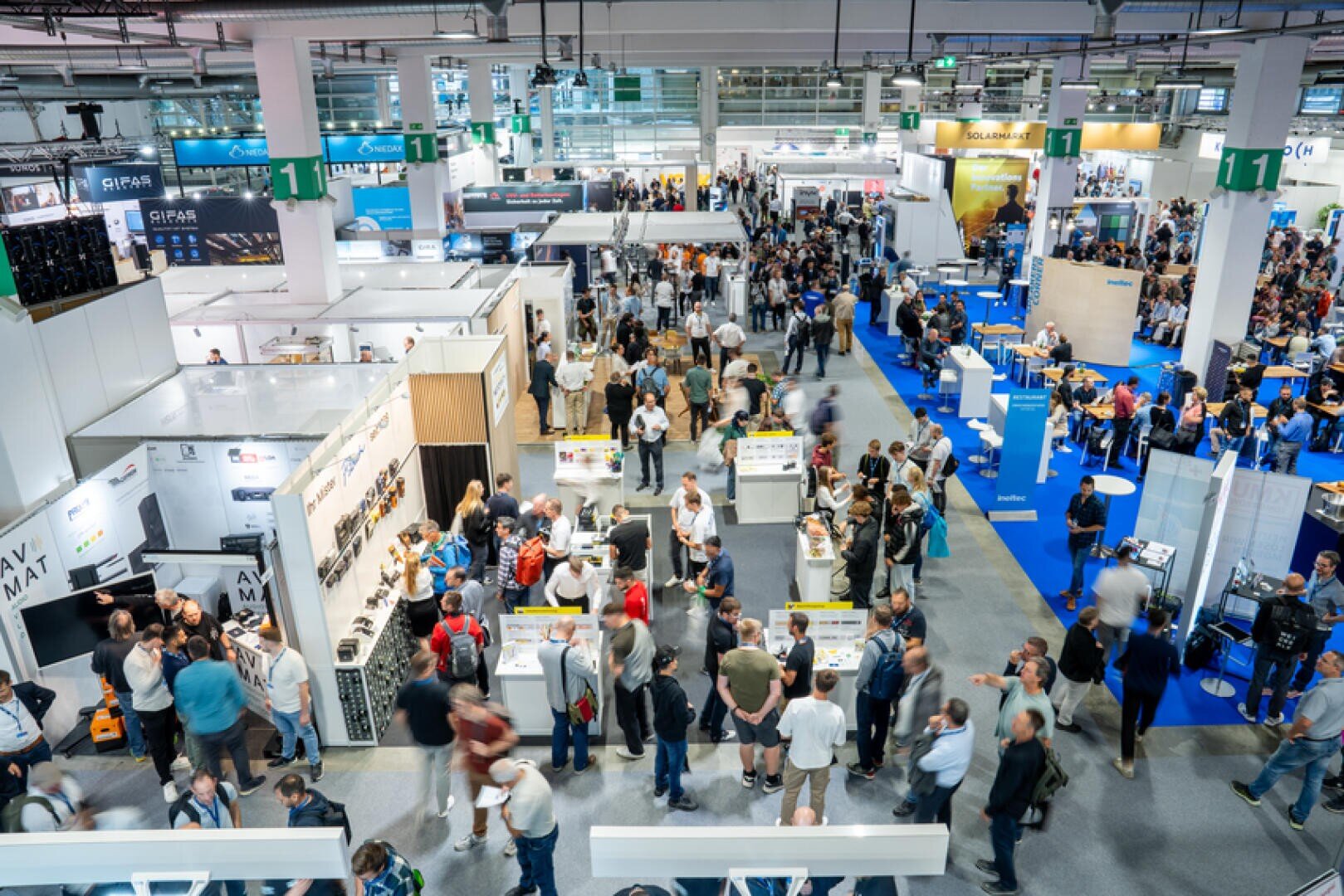 Eine belebte Messe oder ein Kongresszentrum mit vielen Menschen, die zwischen den Ständen und Auslagen umhergehen und mit den Ausstellern ins Gespräch kommen. Der Veranstaltungsraum ist hell beleuchtet, mit Bannern, Schildern und einem blauen Teppichboden.