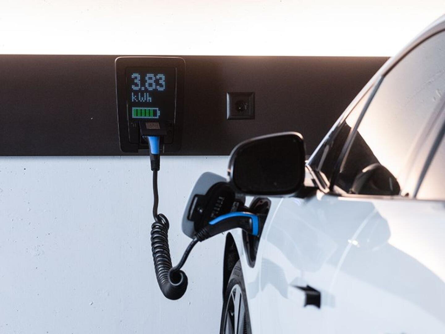 Ein weißes Elektroauto wird an einer Wandladestation geladen, die 3,83 kWh anzeigt. Das Ladekabel ist in das Auto eingesteckt, und die Batterieanzeige zeigt eine Teilladung an.