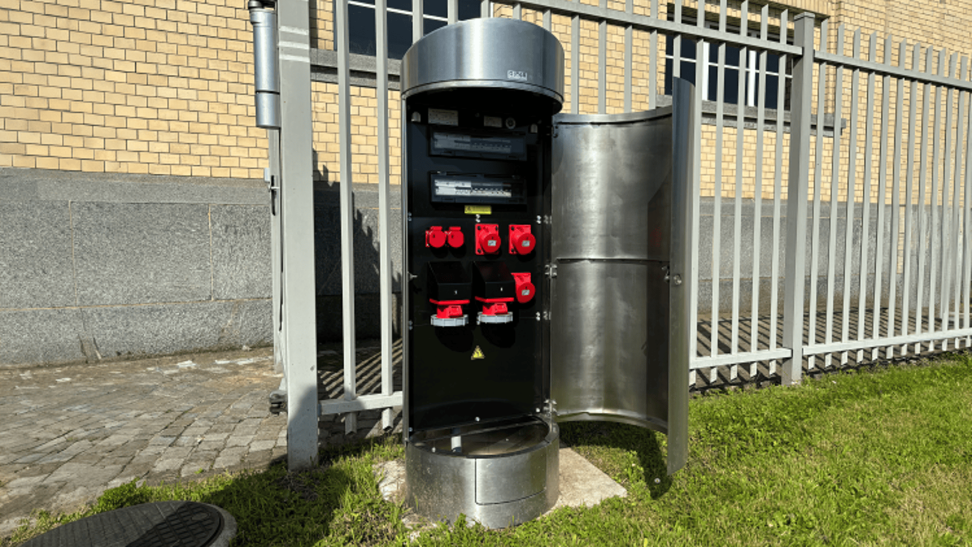 Ein metallischer Schaltschrank für den Außenbereich, durch dessen offene Tür rote und schwarze Schalter und Steckdosen zu sehen sind. Er steht auf dem Gras neben einem Bürgersteig, neben einem grauen Metallzaun und einem Backsteingebäude.