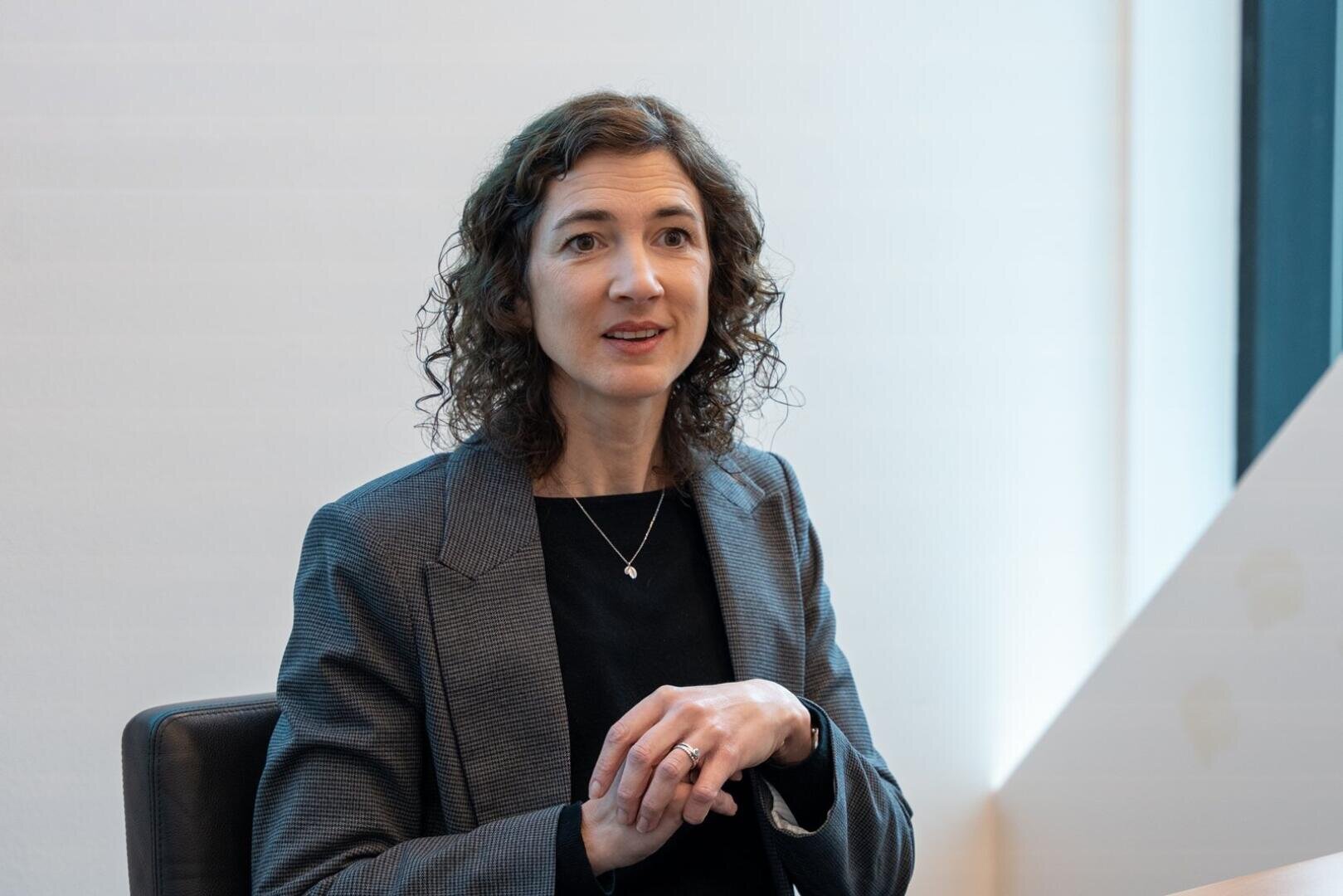 Une femme aux cheveux bruns bouclés, vêtue d'un blazer gris et d'un haut noir, est assise à une table et gesticule avec ses mains tout en parlant dans un bureau moderne et lumineux.
