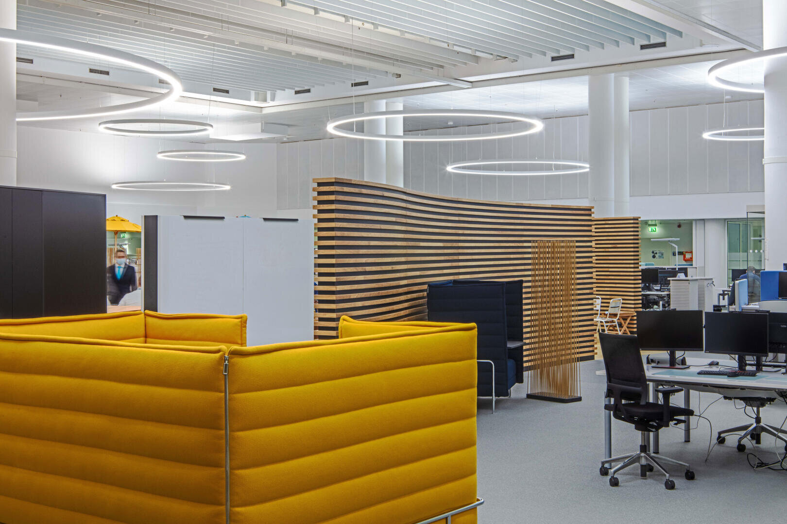 Un espace de bureau ouvert moderne avec des sièges rembourrés jaunes, des chaises de bureau noires, des bureaux avec des ordinateurs, des cloisons de séparation en lattes de bois et de grands plafonniers ronds. L'espace se caractérise par un design minimaliste et moderne.