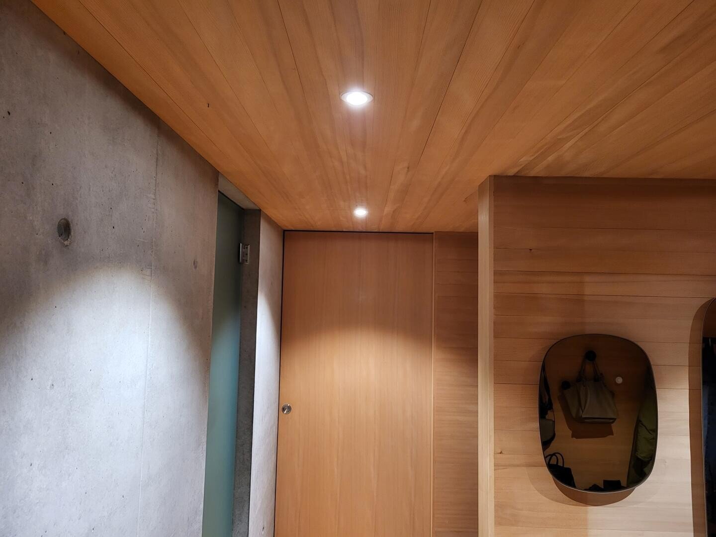 Ein moderner Innenraum mit Holzdecke und -wänden, Einbaulampen, einer geschlossenen Tür, einer Betonwand und einer kleinen Nische, in der eine Tasche hängt.