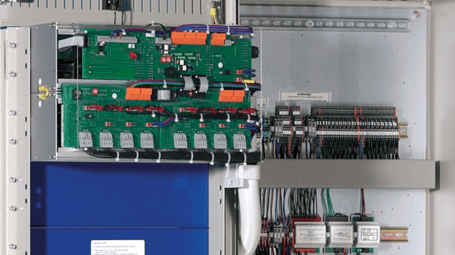 Gros plan d'un panneau de contrôle électrique ouvert montrant des cartes de circuit vertes, du câblage, des connecteurs et des composants électroniques organisés à l'intérieur d'un boîtier métallique.