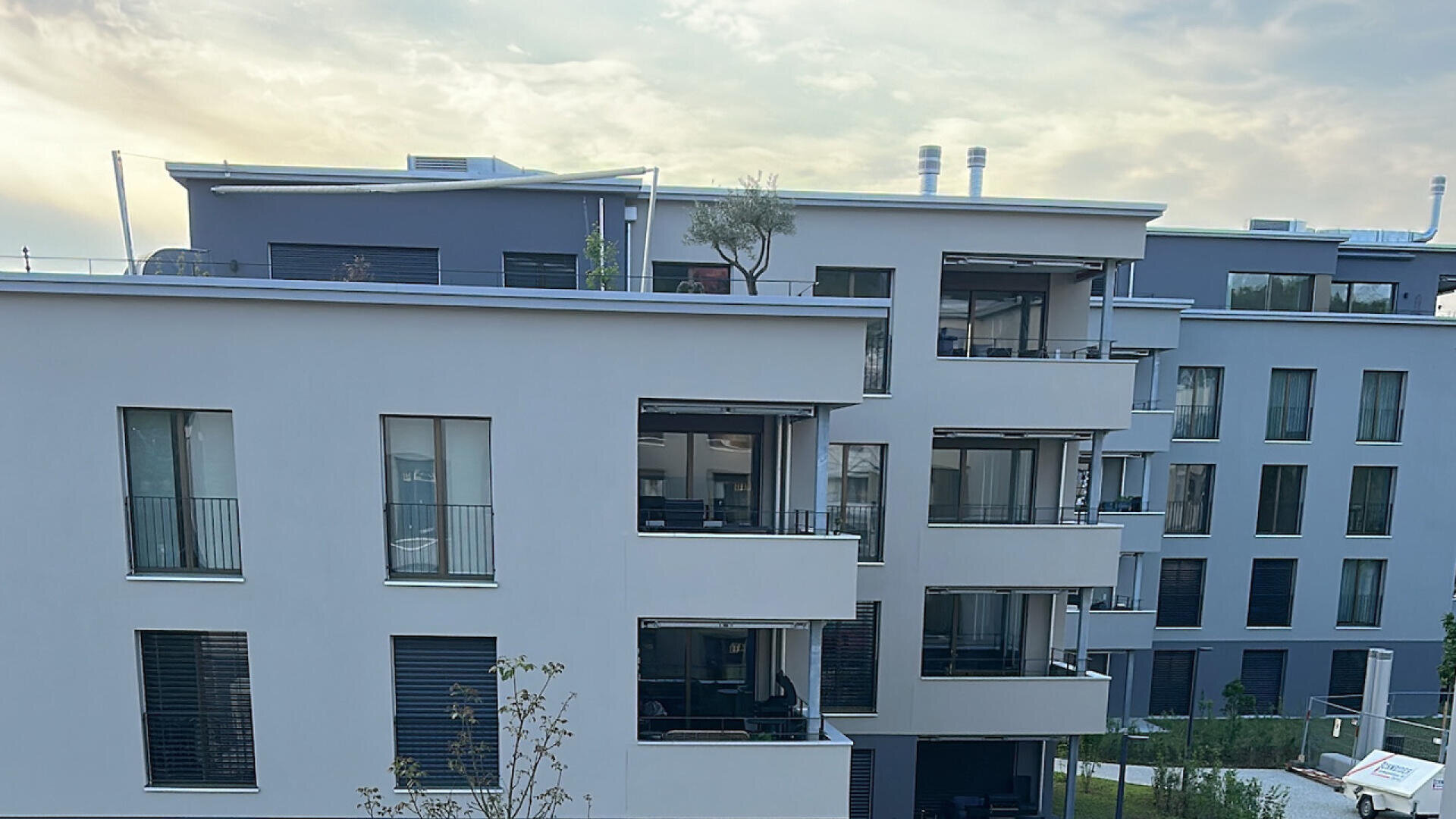 Moderne Apartmentgebäude mit Balkonen, großen Fenstern und Pflanzen auf den Dächern unter einem bewölkten Himmel bei Sonnenuntergang.