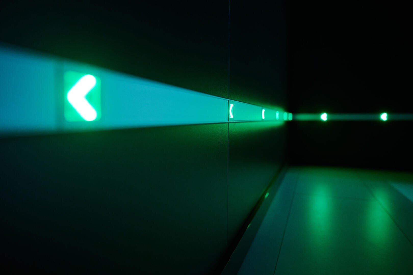 Un couloir sombre avec une série de panneaux fléchés verts lumineux sur le mur, tous orientés vers la gauche, créant une atmosphère futuriste et minimaliste.