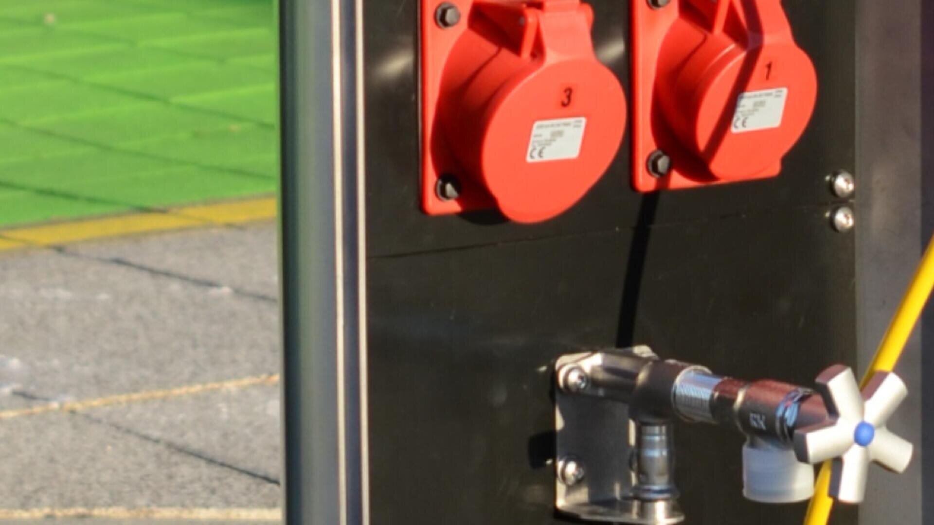 Gros plan de deux prises de courant industrielles rouges sur un panneau métallique noir, avec une vanne connectée et un câble jaune ; un revêtement de sol extérieur vert et gris est visible à l'arrière-plan.
