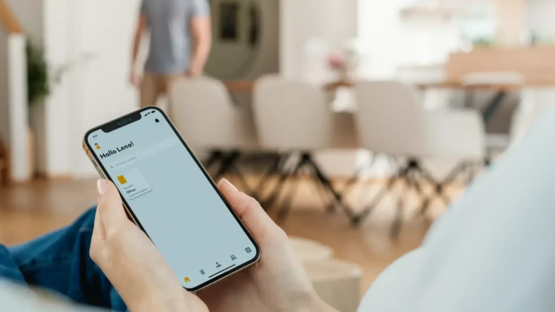 Une personne tenant un smartphone affichant une application de gestion de fichiers, avec un message disant Hello, Lena ! En arrière-plan, une personne floue se tient dans une salle à manger moderne avec des chaises et une table.
