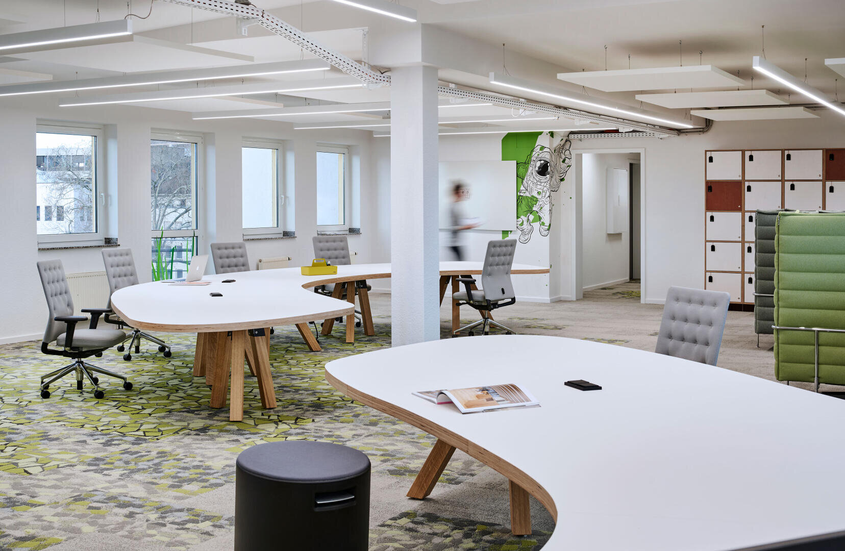 Bureau moderne en open space avec de grands bureaux blancs incurvés, des chaises ergonomiques et une moquette à motifs. Des casiers, des fenêtres et une personne floue qui marche en arrière-plan créent un espace de travail propre et aéré.