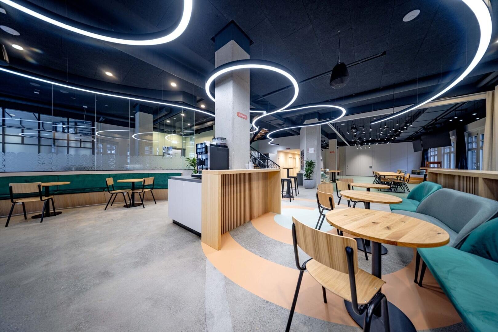 Café moderne et spacieux avec des tables en bois, des chaises, des sièges rembourrés en pétrole et des plafonniers LED ronds uniques. L'espace dispose de grandes fenêtres, d'un comptoir à café et d'un mobilier minimaliste.