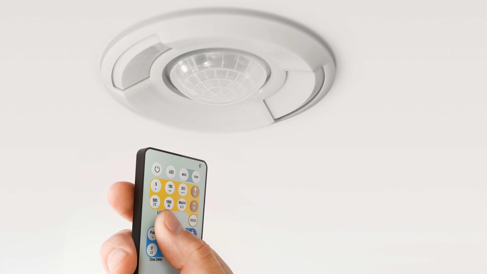 Une main munie d'une télécommande est dirigée vers un capteur de mouvement circulaire monté au plafond.