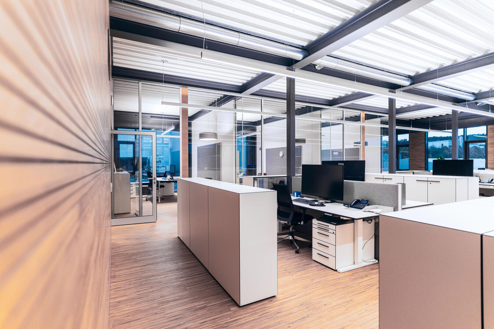 Bureau moderne en open space avec un sol en bois clair, des bureaux blancs, des écrans d'ordinateur, des cloisons en verre et de la lumière naturelle qui pénètre par de grandes fenêtres. L'espace semble propre, organisé et inoccupé.