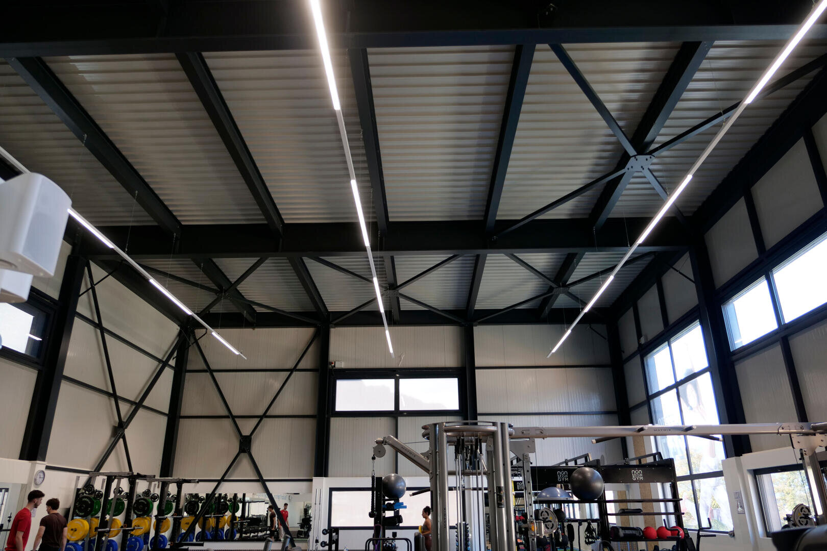 Une salle de sport moderne avec de hauts plafonds, des poutres apparentes, de grandes fenêtres, des supports pour haltères, des appareils de fitness et quelques personnes qui s'entraînent. Des lumières LED brillantes courent sur le plafond.
