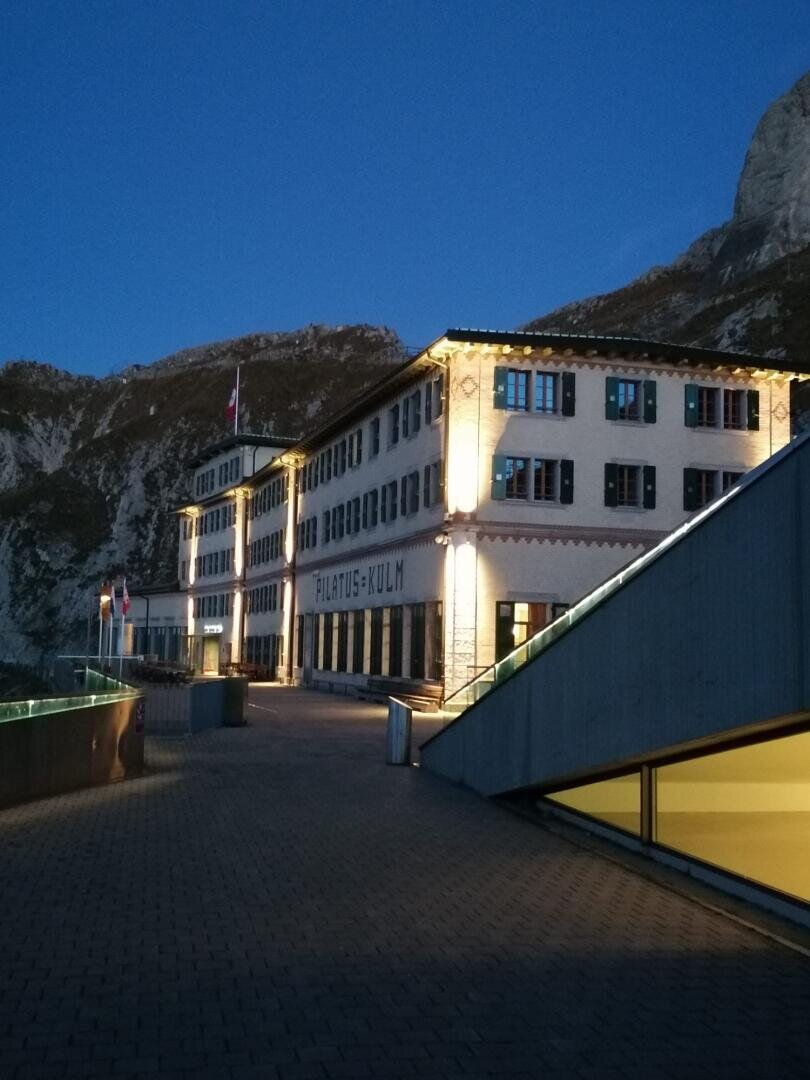 Un grand bâtiment éclairé portant l'inscription "Pilatus-Kulm" se dresse au crépuscule devant un décor de montagne avec un trottoir pavé et des balustrades modernes en verre au premier plan.