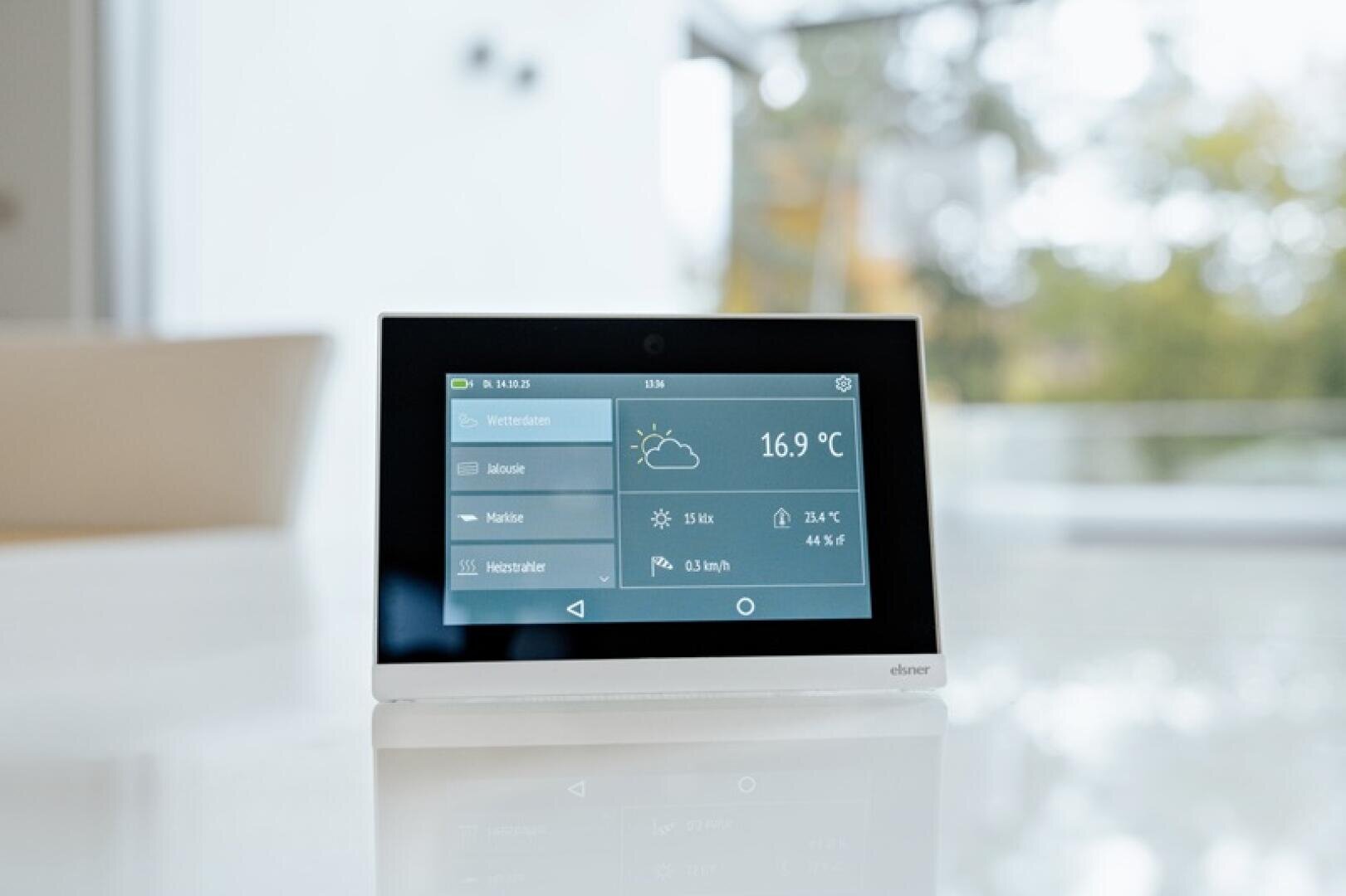 Une station météo numérique affiche sur un écran tactile une température de 16,9 °C, les conditions météorologiques et d'autres informations. L'appareil est posé sur une surface blanche dans une pièce lumineuse et moderne avec de grandes fenêtres et de la verdure floue à l'extérieur.