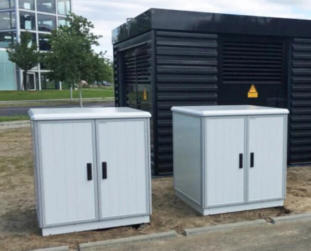 Zwei hellgraue Stromkästen mit Doppeltüren stehen auf einem Betonsockel im Freien vor einem größeren schwarzen Industriegebäude. Im Hintergrund sind Bäume und ein modernes Glasgebäude zu sehen.