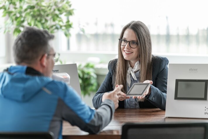Eine Frau in Geschäftskleidung lächelt, während sie einem Mann am anderen Ende des Tisches in einem hellen Büro mit Produktauslagen und Grünpflanzen im Hintergrund ein digitales Tablet zeigt.