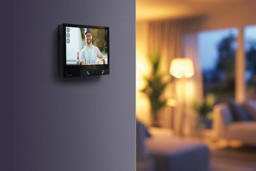 Un écran d'interphone vidéo sur un mur montre un livreur souriant. L'arrière-plan révèle un salon moderne à la lumière tamisée, avec une lampe, un canapé et des rideaux, légèrement flous.