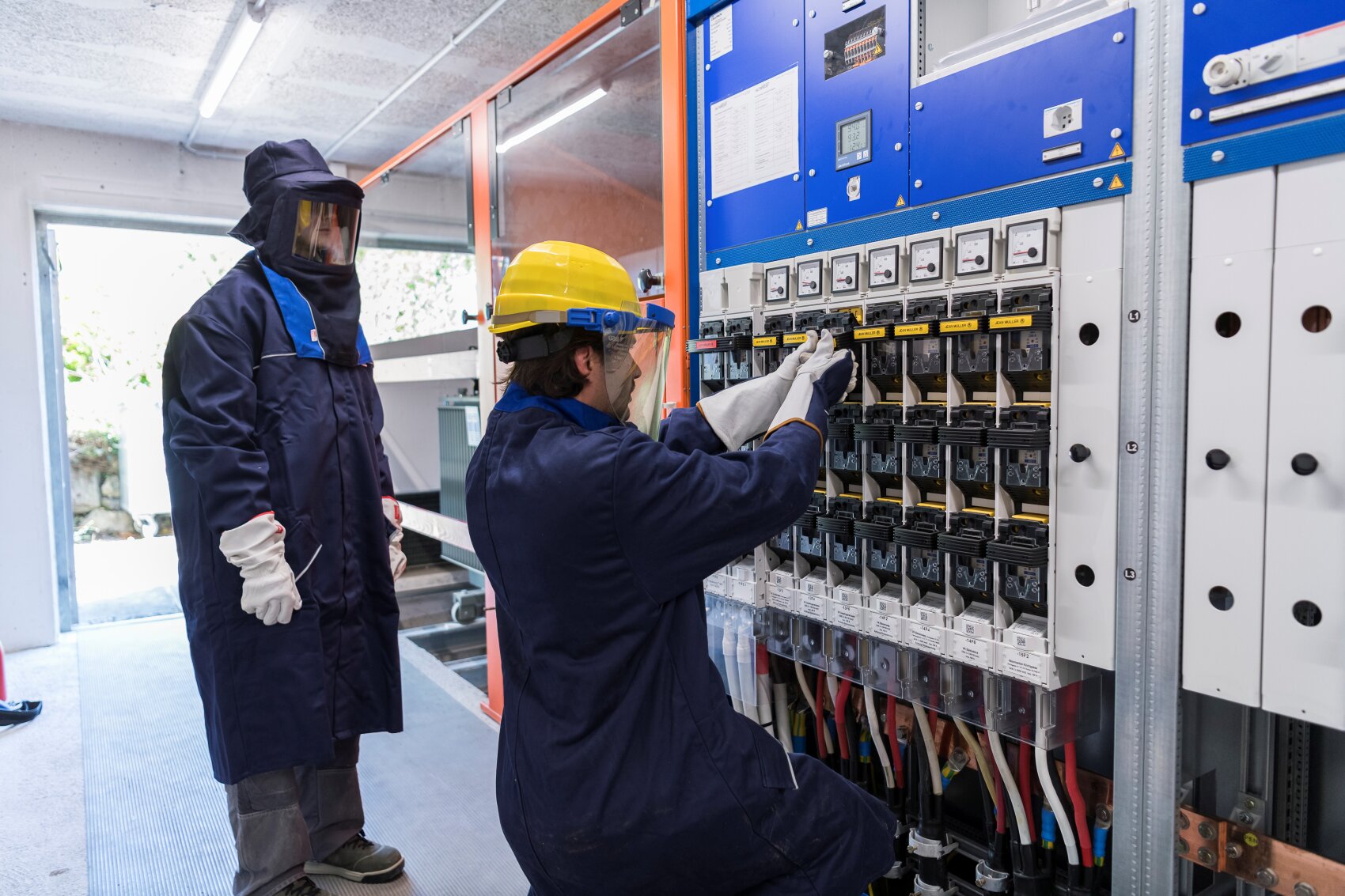 Zwei Arbeiter in Schutzkleidung und mit Helm überprüfen und bedienen elektrische Schalter auf einer großen Schalttafel in einer Industrieanlage. Ein Arbeiter kniet und stellt die Schalter ein, während der andere in der Nähe steht.